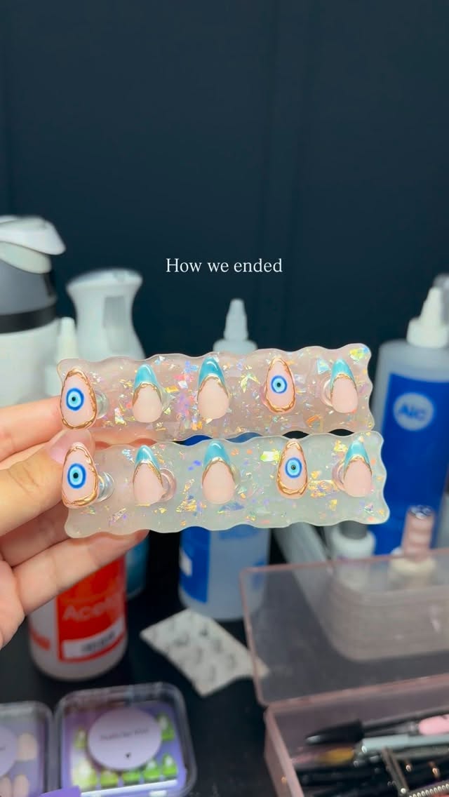 Nude press-ons with gold rims, baby-blue accents, and evil-eye details on a glitter stand.