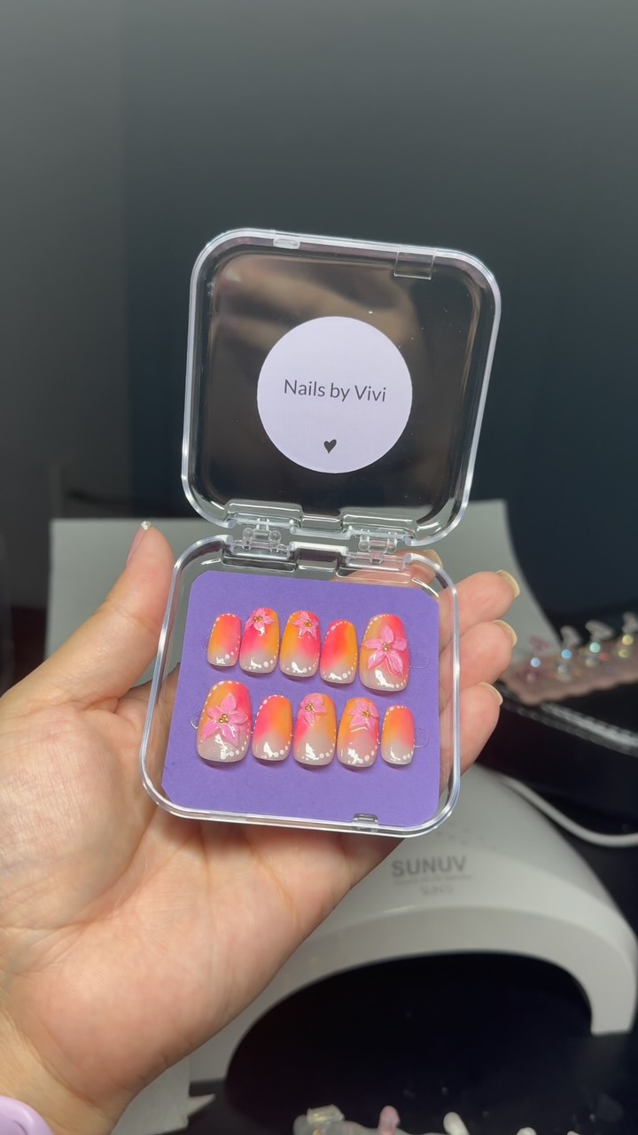 Pink-to-orange sunset French press-ons with tiny flowers, inside a clear Nails by Vivi case.